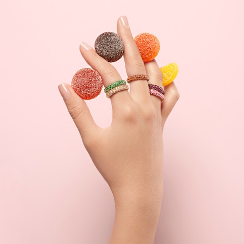A hand with rings from Maison De Greef holding colorful candies between each finger, in front of a pale pink background